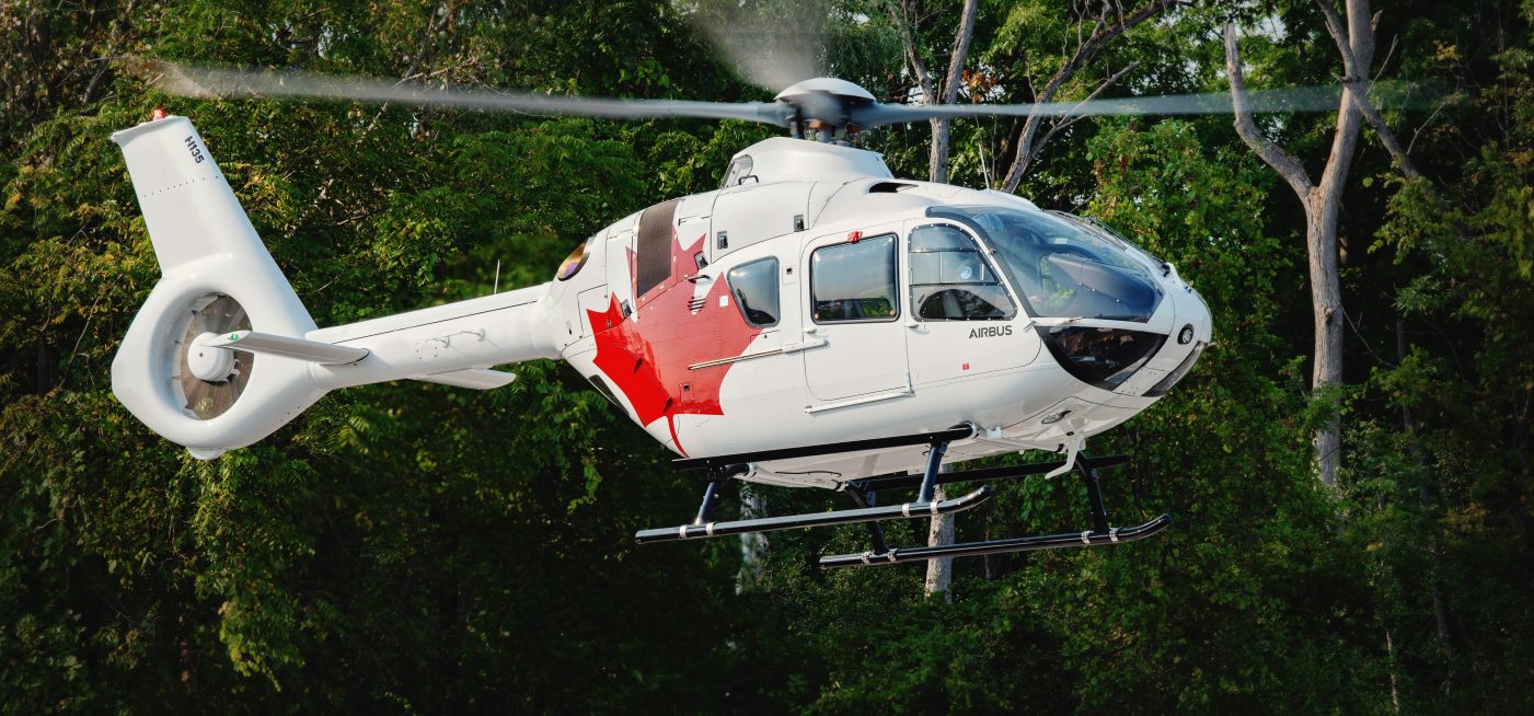 White H135 helicopter with red maple leaf hovers with green trees in the background