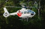 White H135 helicopter with red maple leaf hovers with green trees in the background