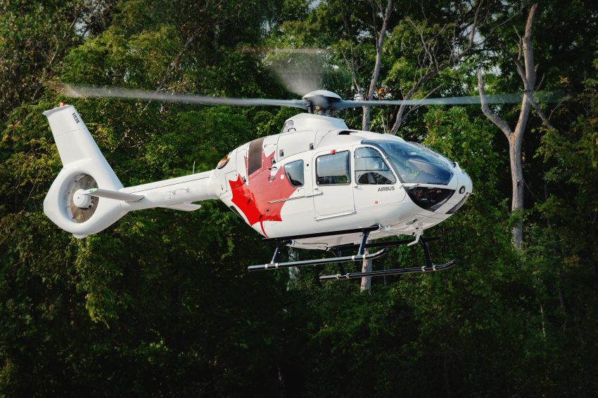 White H135 helicopter with red maple leaf hovers with green trees in the background